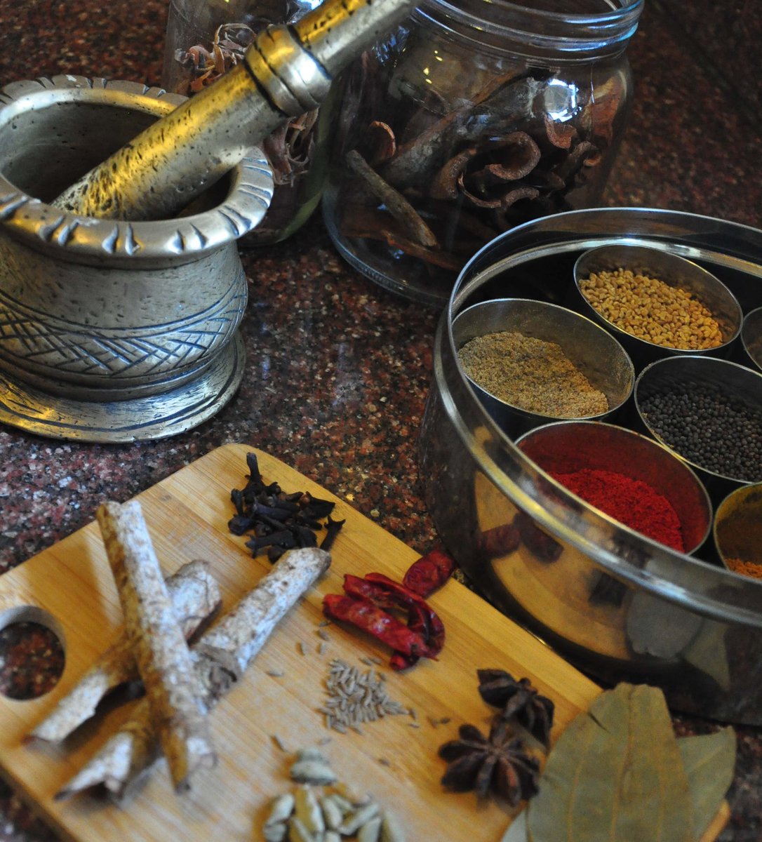 A brass mortar and pestle beside a stainless steel masala dabba and whole spices — star anise, cloves, cinnamon, cardamom, bay leaf, dried chillies — laid out on a wooden board